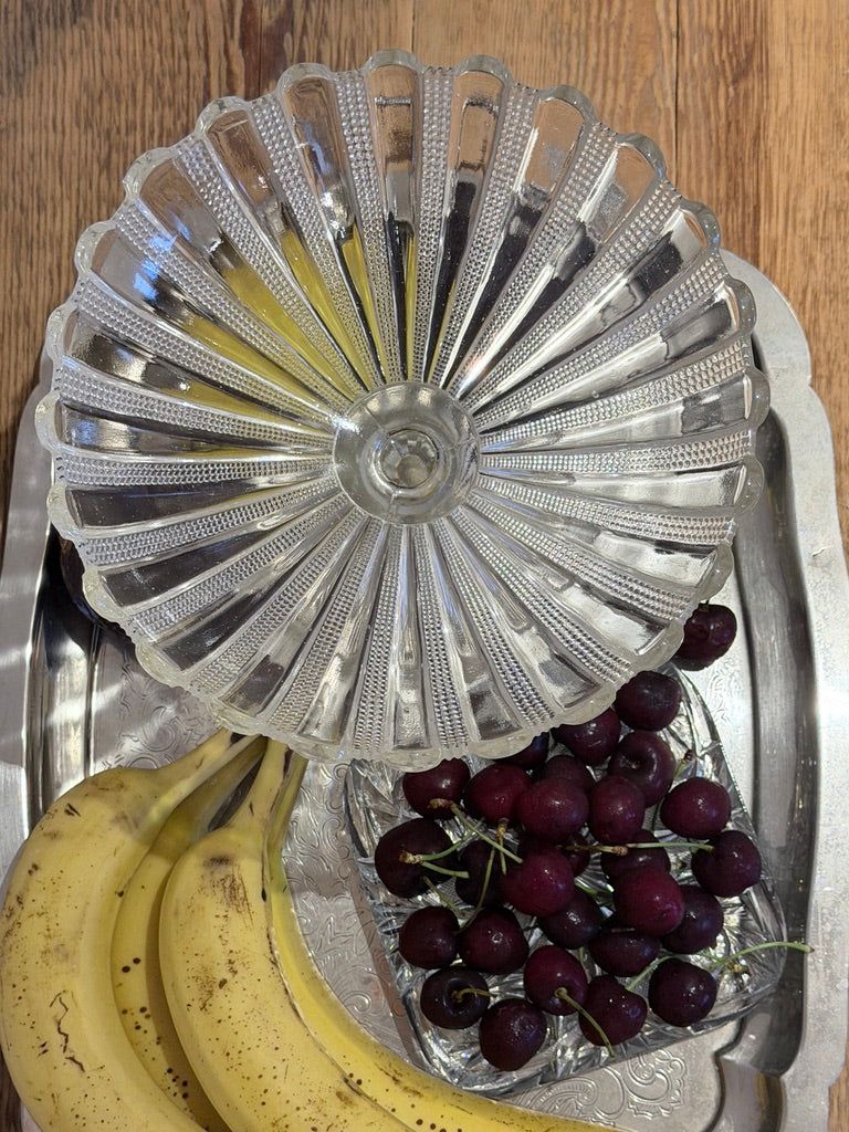 A vintage clear glass pedestal bowl with a scalloped rim and textured ribs, shown on a silver tray with bananas and cherries.