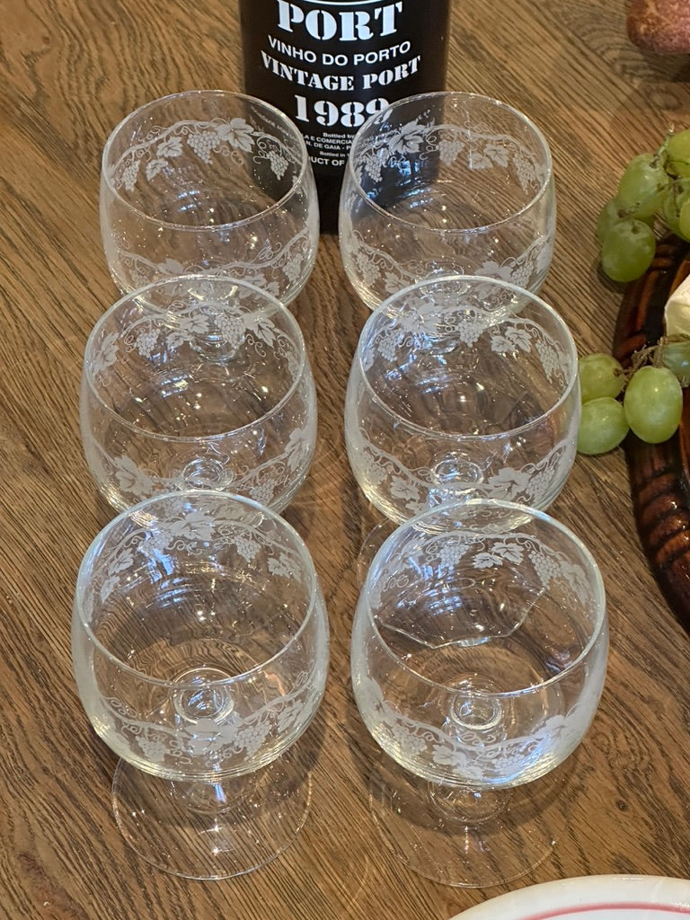 Six clear crystal port glasses with white floral engravings arranged on a wooden table next to a bottle of vintage Port wine and green grapes.