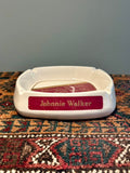 A white ceramic ashtray with a red front plaque reading 'Johnnie Walker' in gold lettering
